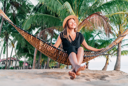 Positive young woman sitting in hammock on the tropical palm beachの写真素材