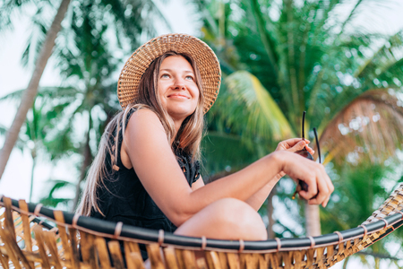 Happy smiling young woman in straw hat sitting in hammock with palm trees backgroundの写真素材