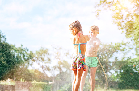 Brother sptays his slittle sister water from garden hose. Children have fun in hot summer dayの写真素材