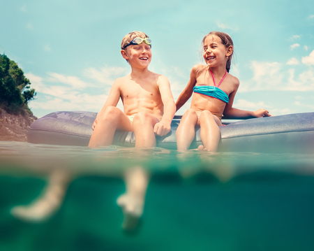Sister and brother sitting on inflatable mattress and enjoying the sea water, cheerfully laughing when swim in the sea. Careless childhood time unusually water level shot image.の写真素材