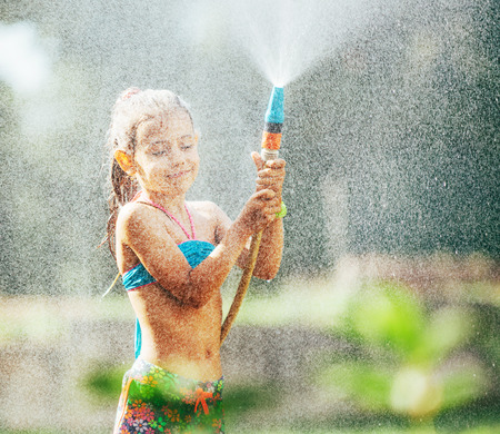 Cute little girl sprinkls a water for herself from the hose, makes a rain. pleasure for hot summer daysの写真素材