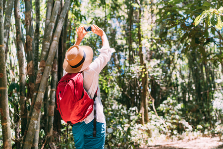 Woman with backpack on trek through jungle forest stopping taking picture with smartphoneの写真素材