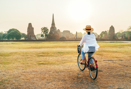 Young female weared light summer clothes and hat riding bicycle near Ayutthaya historical park in Thailand covered with morning sunlightの写真素材