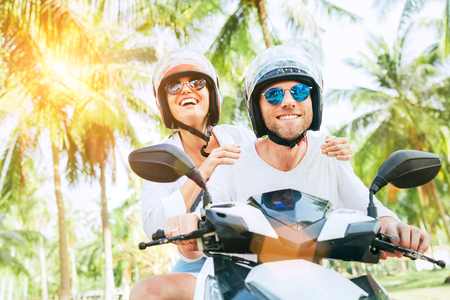 Happy smiling couple travelers riding motorbike scooter in safety helmets during tropical vacation under palm treesの写真素材
