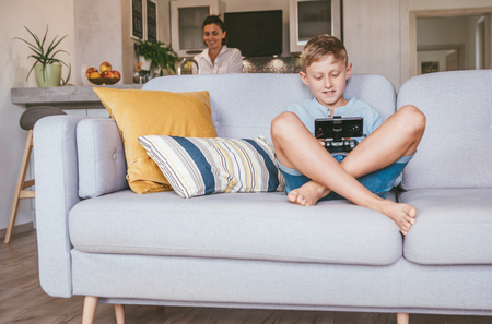 Boy plays with electronic device. His mother prepared something in the blurred  kitchenの写真素材