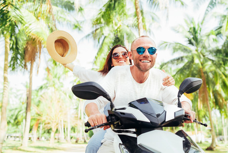 Laughing happy couple travelers riding motorbike during their tropical vacation under palm trees. Woman raised hand with hat up.の写真素材