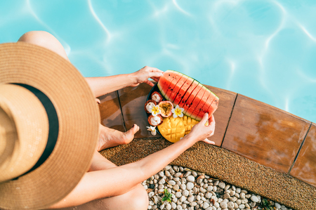 Caucasian Woman in straw hat sitting on swimming pool side near plate of tropical fruits- camera and trying to eat watermelon. Top view oimage.の写真素材