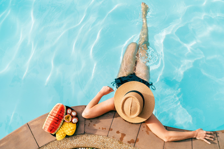 Top view of Woman in straw hat sitting on swimming pool side near plate of tropical fruits- cameraの写真素材