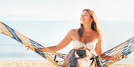 Happy smiling female swinging in hammock hinged between palm trees at the sea sideの写真素材