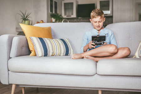 Preteen boy losted in computer game. Sits at home on coze sofa, uses gamepad with smartphoneの写真素材