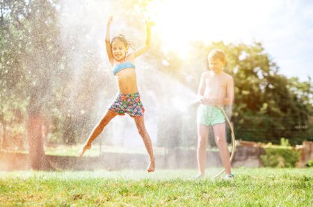 Happy little girl jumps under water, when brother pours her from garden hose. Hot summer days activity.の写真素材