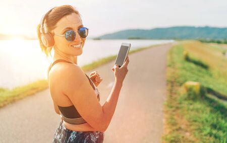 Young beautiful female starting jogging and listening to music using smartphone and  wireless headphones cheerfully smiling in cameraの写真素材