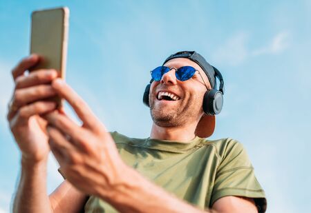 Bearded man in baseball cap, wireless headphones and blue sunglasses found something funny in smartphone device and cheerful laughing.の写真素材