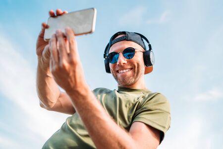 Bearded man in baseball cap, wireless headphones and blue sunglasses cheerful smiling taking a selfie picture using modern smartphone device.の写真素材