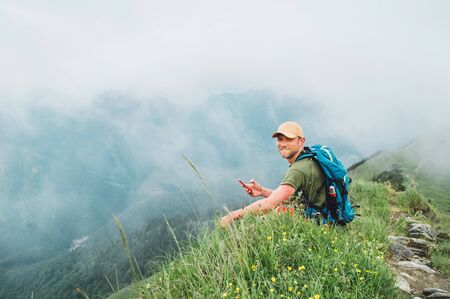 Tired backpacker man browsing internet using smartphone enjoying cloudy valley bottom walking by the foggy cloudy weather mountain rangeの写真素材