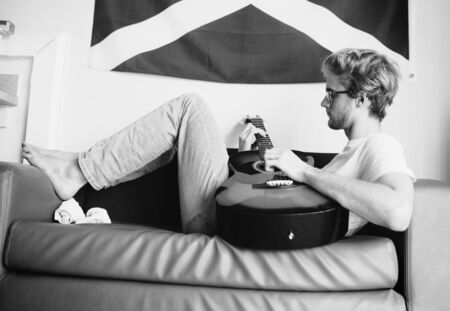Black and white vintage image style middle shot of young man lying on sofa and playing on guitar in teen room.の写真素材