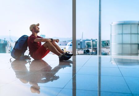Alone backpacker traveller sitting on the airport terminal floor and waiting for boarding at aircraft which prepared for flight behind a windowの写真素材