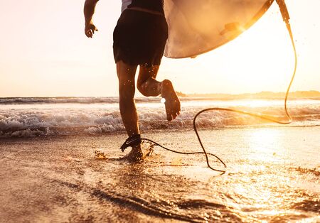Young man Surfer taking surfboard and running with long surf board to waves on the evening sunset sky background. Active holidays spending concept.の写真素材