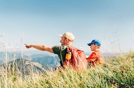 Father showing something interesting to his teenager son sitting on the grass during their mounting hiking walking.の写真素材