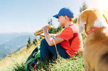 Little smiling boy weared baseball cap enjoying a huge baguette sandwich and his beagle dog friend watching it during a mountain hiking resting snack break time.の写真素材