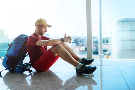 Alone backpacker traveller sitting on the airport terminal floor and waiting for boarding at aircraft which prepared for flight behind a windowの写真素材