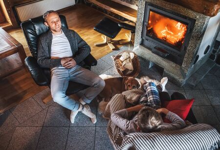 Father with son sitting in comfortable armchairs in their cozy country house near fireplace and enjoying a warm atmosphere and flame moves. Their beagle friend dog sitting beside on white sheepskin.の写真素材