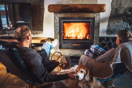Father with son sitting in comfortable armchairs in their cozy country house near fireplace and enjoying a warm atmosphere and flame moves. Their beagle friend dog sitting beside on white sheepskin.の写真素材