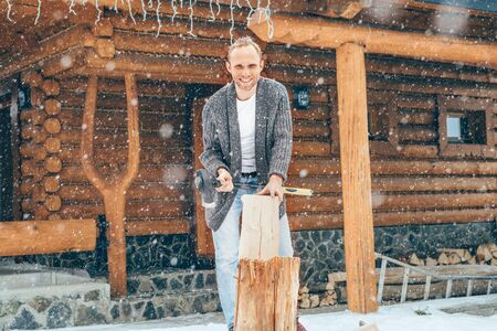 Man chopping wood on snowy yard for a house fireplace with heavy snowflakes background . Winter countryside holidays concept imageの写真素材