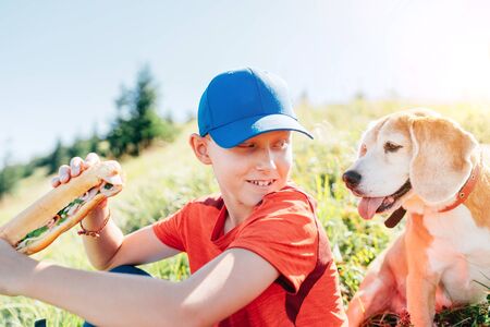 Little smiling boy weared baseball cap with a huge baguette sandwich with his beagle dog friend during a mountain hiking resting snack break time.の写真素材
