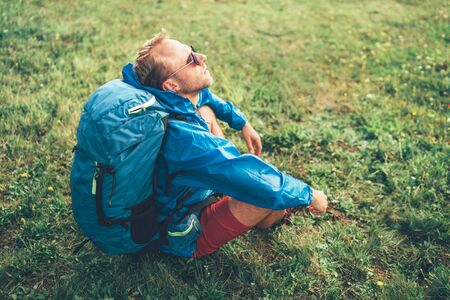 Backpacker man has a rest break sitting on green grass and enjoying mountain walking, he raised his head into air.の写真素材