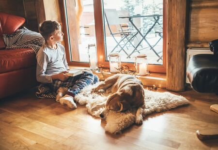 Boy reading book on the floor near slipping his beagle dog on sheepskin in cozy home atmosphere.の写真素材