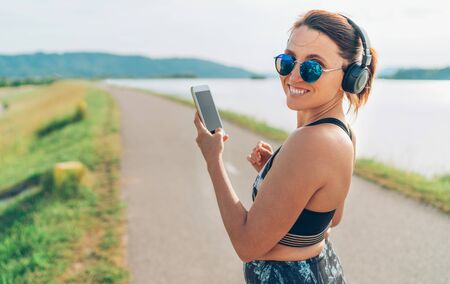 Young beautiful female listening to music using smartphone and  wireless headphones and starting jogging and cheerfully smiling in cameraの写真素材