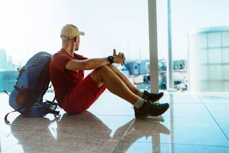 Alone backpacker traveler sitting on the airport terminal floor, looking at hall window and waiting for boarding at aircraft which prepared for flight behind a windowの写真素材