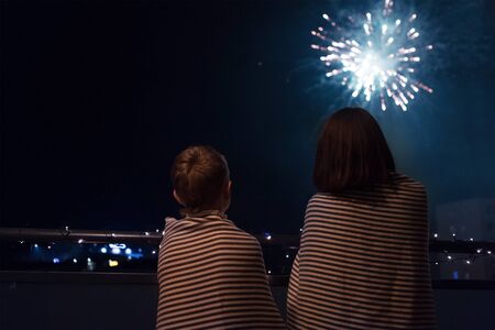 Mother and son looking at New Year celebration fireworks in night sky warmly wrapped in striped plaids standing on home balcony.の写真素材