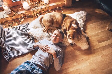 Boy lying on the floor and near beagle dog sleeping on sheepskin in cozy home atmosphere. Peaceful moments of cozy home, holiday time concept imageの写真素材