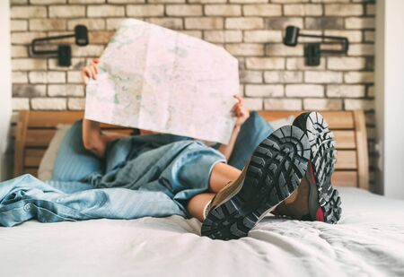 Young female inspecting a city map resting on the bed under the blanket weared trekking boots on bed...