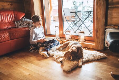 Boy reading book on the floor near slipping his beagle dog on sheepskin in cozy home atmosphere. Peaceful moments of cozy home concept image.の写真素材