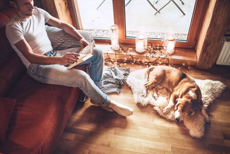 Man reads book sitting on the cozy couch and his beagle dog sleeps on sheepskin on floor. Calm and cozy home atmosphere.の写真素材