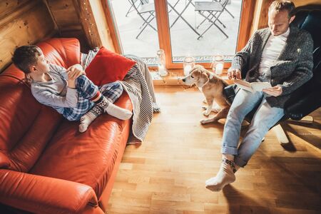 Father, son and his beagle dog spending holiday time in cozy country house. Dad reading a book, boy sitting and listening, dog looking in window. Winter family vacationの写真素材