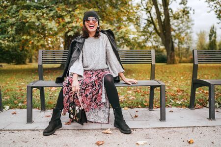 Sincerely laughing female dressed fashion boho style colorful long dress, warm knitted sweater with black leather biker jacket and Beanie Hat with flap bag sitting on bench at autumn city parkの写真素材