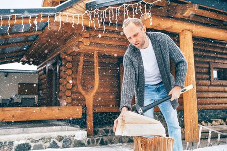 Man chopping wood on snowy yard for a house fireplace with heavy snowflakes background . Winter countryside holidays concept imageの写真素材