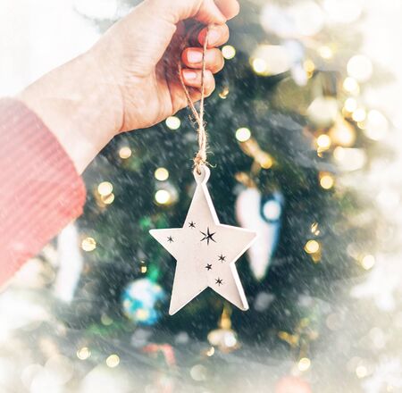 Close up image of female hand hanging Christmas wooden star decoration on spruce tree. Unfocused green branches bokeh with new year lights on the background.の写真素材