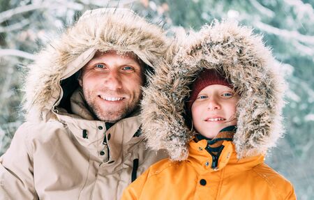 Father and son dressed in Warm Hooded Casual Parka Jacket Outerwear walking in snowy forest cheerful smiling faces portrait. Father and son relatives and winter outfit concept image.の写真素材