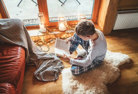 Boy reading book on the floor on sheepskin in cozy home atmosphere. Peaceful moments of cozy home concept image.の写真素材