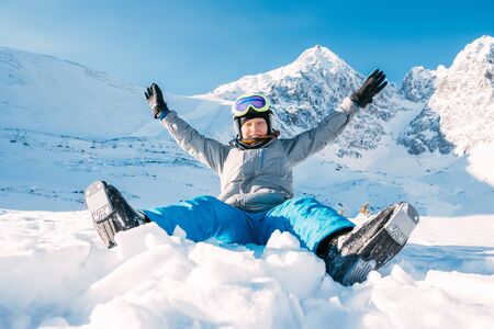 Portrait of laughing boy sitting on the snowy slope, rising an arms up with High Tatra Mountains range background. Carefree childhood winter holidays concept image.の写真素材