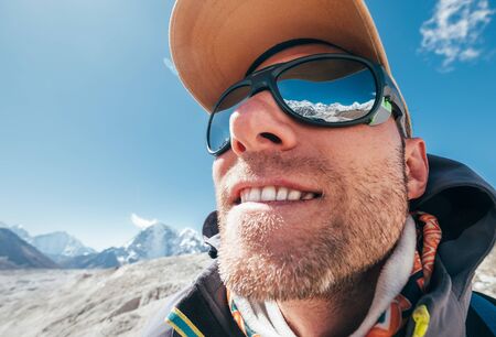 Ultra-wide lens angle portrait shot of high altitude mountain smiling unshaven happy hiker in baseball cap with snow peaks and mountain range beside him and reflected in mirror UV protected sunglassesの写真素材