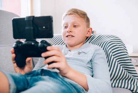Preteen boy sits at home on cozy sofa, uses game pad with smartphone.の写真素材