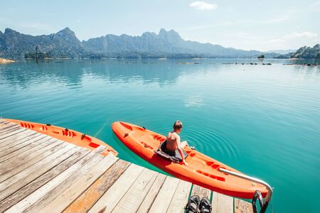 Little boy sitting on orange kayak near the pier and dangling feet making radial waves on Cheow Lan Lake, Khao Sok national park, Thailandの写真素材