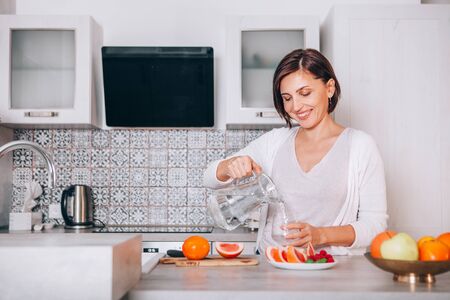 Young sincerely smiling female pouring a purified mineral water into transparent glass decanter at modern kitchen. Plenty of apples, grapefruits, kaki and oranges fruits, berries are on the tables. の写真素材