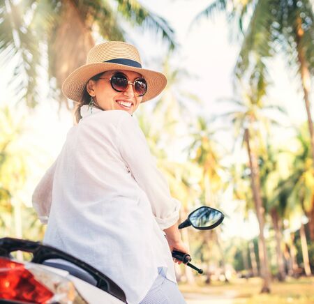 Happy smiling woman in straw hat and sunglasses riding motorbike under palm treeの写真素材